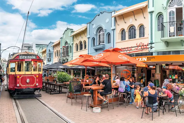 A red heritage City Tour tram passing pastel art-deco shopfronts and orange café umbrellas on New Regent Street, Christchurch, with people seated at outdoor tables.