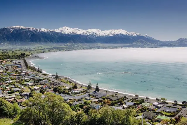 Kaikoura bay seen from above — the snow-capped Seaward Kaikoura range behind a curved shoreline, sea fog lying flat across turquoise water.